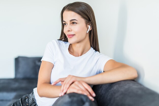 Cute smiling woman lying on couch while listening via airpods to music in bright living room