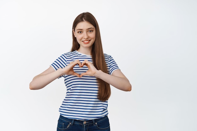 Cute smiling woman showing heart sign near chest, standing over white background. Relationship and romance concept