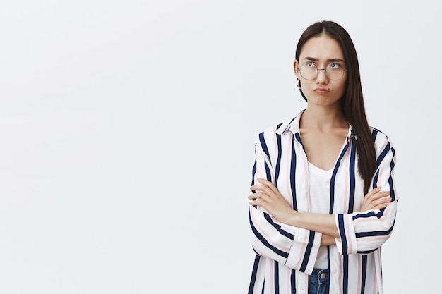 Cute sulking woman with dark hair in glasses and striped shirt, holding hands crossed, pursing lips and staring gloomy at upper left corner