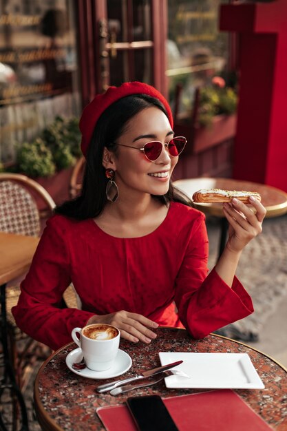 Cute tanned brunette woman in stylish red dress, beret and sunglasses sits in cafe