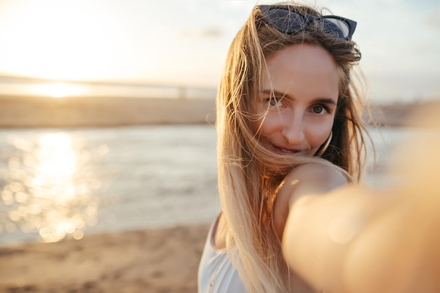 Cute tanned girl with straight hair making selfie on nature background Goodlooking woman in sunglasses taking picture of herself while standing at sea coast early in evening