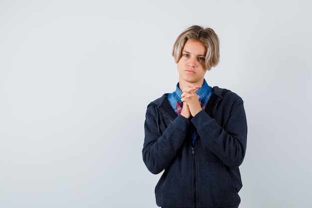 Cute teen boy clasping hands in praying gesture in shirt, hoodie and looking hopeful , front view.