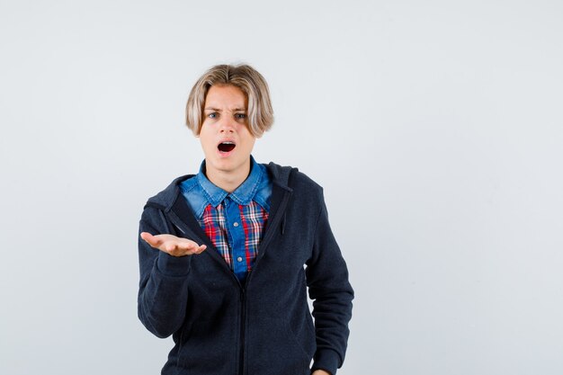 Cute teen boy spreading palm at front in shirt, hoodie and looking bewildered. front view.