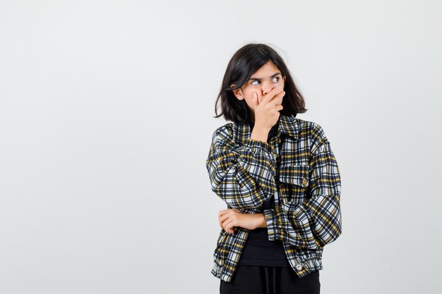 Cute teen girl holding hand on mouth, looking up in checked shirt and looking anxious. front view.