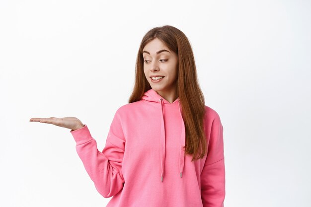 Cute teen girl student, holding copyspace on palm, display product in her hand against white wall, smiling and looking at object, white wall