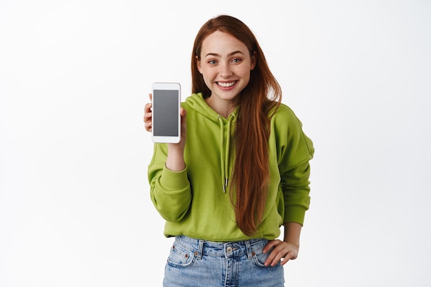 Cute teenage redhead girl, show mobile phone app and smiling happy, introduce smartphone application interface, standing against white background