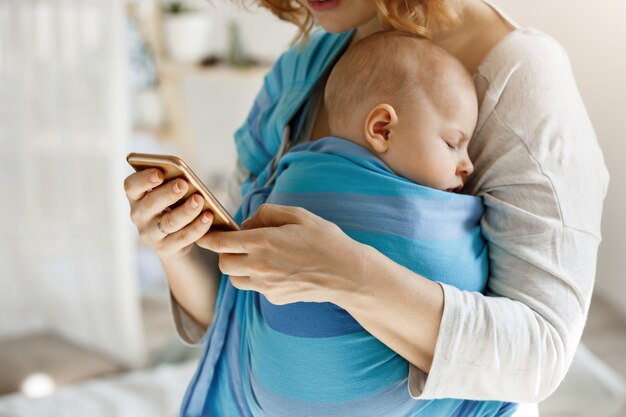 Cute tiny kid napping peacefully while mother hugging him and texting husband by phone asking buy some baby food and diapers. Lifestyle, family concept.