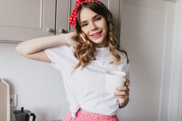 Cute white girl in casual t-shirt standing in kitchen with cup of tea. Indoor shot of relaxed female model drinking coffee in morning.