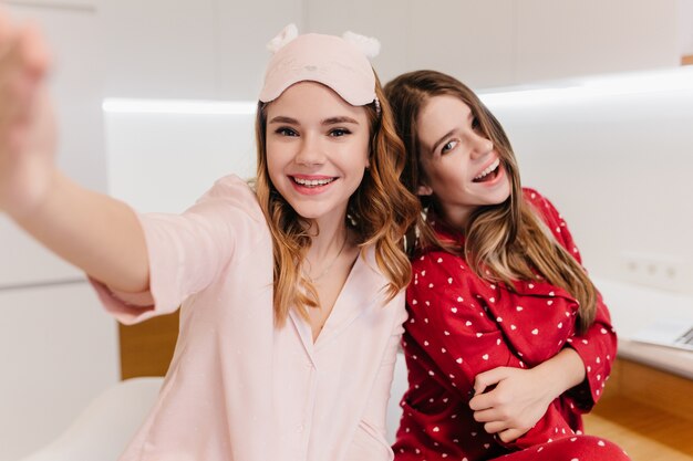 Cute white girl with lovely smile making selfie in light room. Indoor shot of two amazing european ladies enjoying weekend.