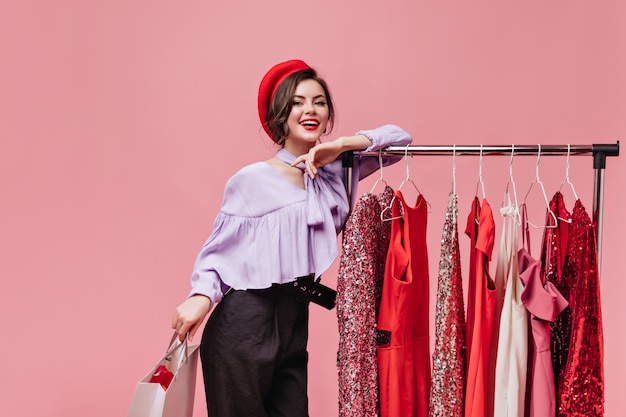 Free Photo cute woman in bright hat and purple blouse is leaning on stand with dresses and posing with package on isolated background.