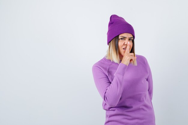 Cute woman showing silence gesture in sweater, beanie and looking sensible , front view.