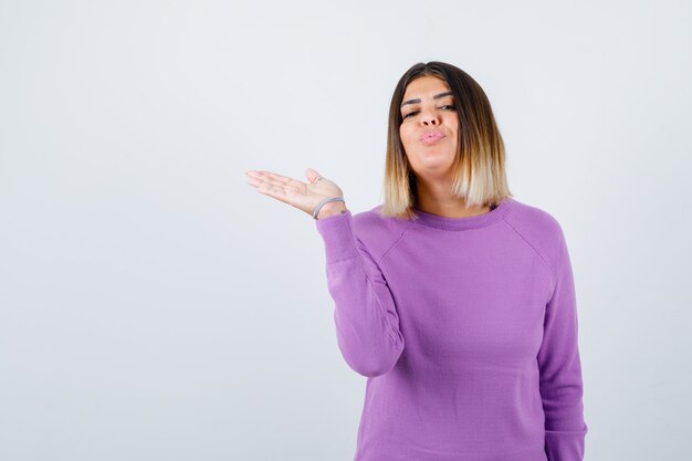 Cute woman spreading palm aside, pouting lips in purple sweater and looking peaceful , front view.