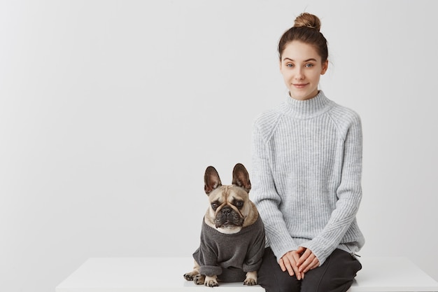 Free photo cute woman with fashion hairstyle and her puppy french bulldog dressed in jumper. female model sitting on table with dog over white wall. friendship concept, copy space