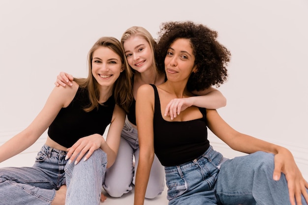 Cute young caucasian and african ladies in tops and jeans look at camera on white background Diverse ethnicity lifestyle