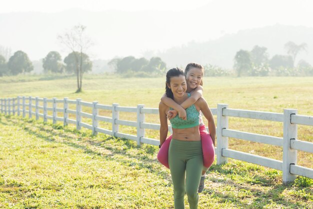 Cute young daughter on a piggy back ride with mother her spending time in countryside. happy family on meadow in summer in nature.  outdoor sports and fitness, exercise learning for kid development.