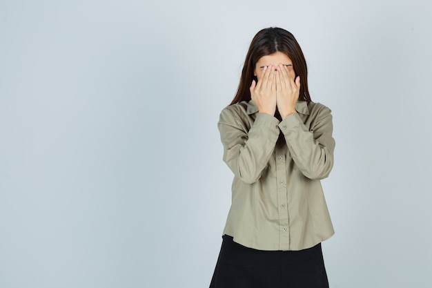 Cute young female in shirt, skirt covering face with hands and looking depressed , front view.