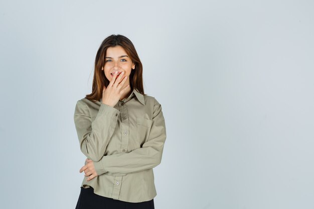 Cute young female in shirt, skirt holding hand on mouth and looking joyful , front view.