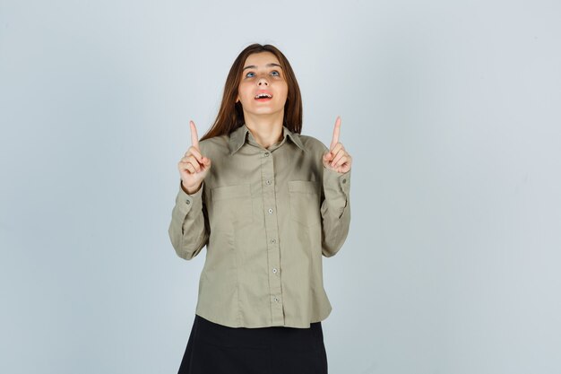 Cute young female in shirt, skirt pointing up and looking hopeful , front view.