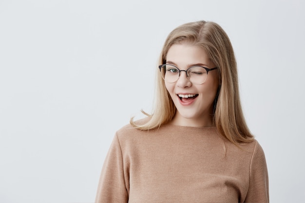 Cute young female with blonde straight hair wearing brown loose sweater and eyeglasses blinking her eyes with pleasure having happy expression. Facial expressions and people emotions concept