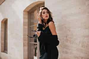 Free photo cute young girl with dark wavy hairstyle and bright makeup, silk dress, black jacket, holding sunglasses in hands and looking away against beige building wall