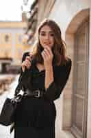 Free photo cute young girl with medium wavy hairstyle, modern makeup, green silk dress, black jacket and stylish accessories posing on daylight street and looking at front