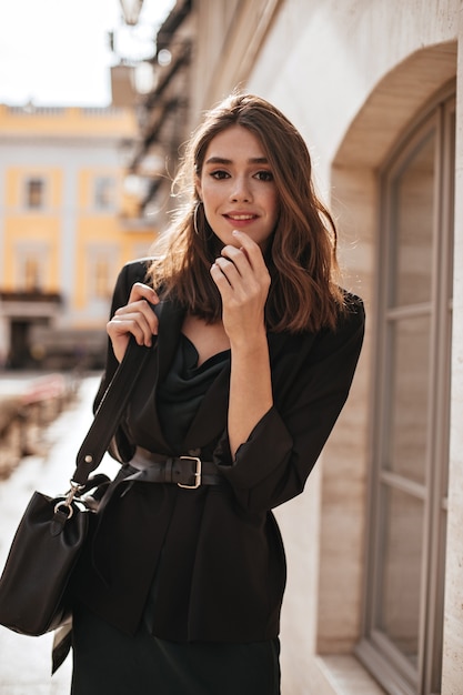 Cute young girl with medium wavy hairstyle, modern makeup, green silk dress, black jacket and stylish accessories posing on daylight street and looking at front