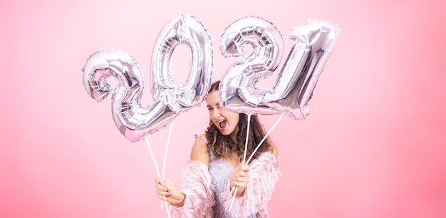 Free photo cute young girl with a smile in a festive outfit posing against a pink studio background and holding silver balloons for the new year concept