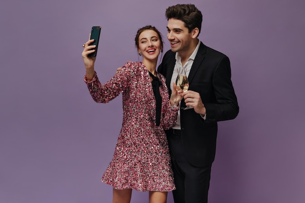 Free photo cute young people in party outfit smiling holding glasses with champagne and video chatting pair posing isolated on violet background