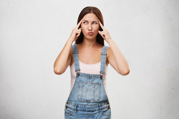 Cute young woman in denim jumpsuit holding fingers at her temples and looking sideways with concentrated serious expression while searching for solution to personal trouble or problems at work