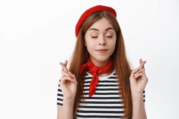 Free Photo cute young woman in fancy hat, close eyes and cross fingers for good luck, making wish, hope for something happen, praying, standing over white wall