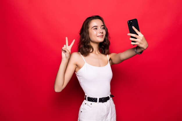 Cute young woman having fun and taking selfie with gesturing peace sign isolated over red 