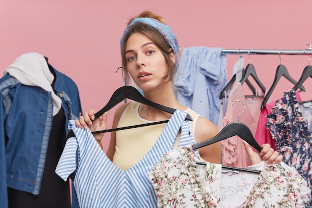 Cute young woman holding two different summer dresses deciding which one is more suitable to wear for a walk. People, clothing, style and fashion