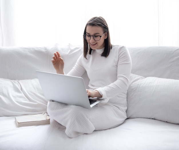 A cute young woman is sitting at home on a white sofa in a white dress and working on a laptop.