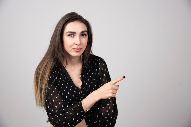 Cute young woman pointing a finger away on a gray wall.