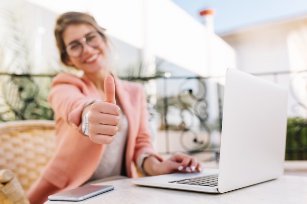 Free Photo cute young woman, student, business lady showing thumbs up, well done, sitting in outdoor cafe on terrace with laptop. wearing pink smart clothes.