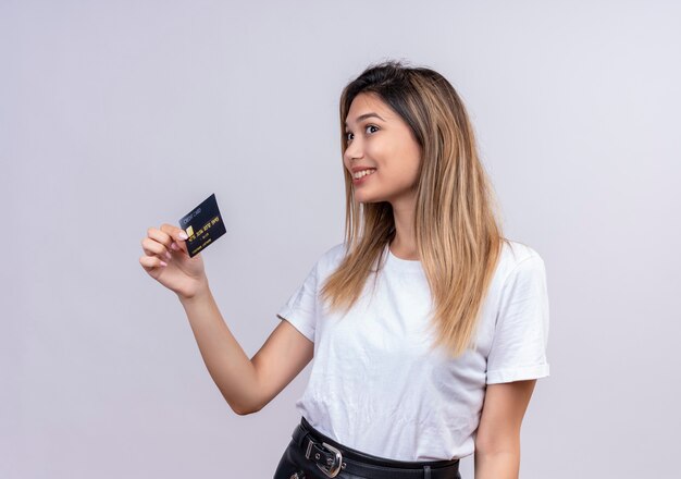 A cute young woman in white t-shirt smiling and showing credit card on a white wall