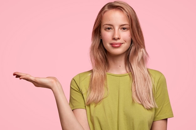 Cute young woman with pleasant appearance, raises palm, advertises something, dressed in green casual t shirt, stands against pink wall