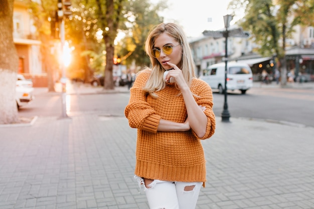 Cute young woman with shiny blonde hair posing in city square with pleased face expression