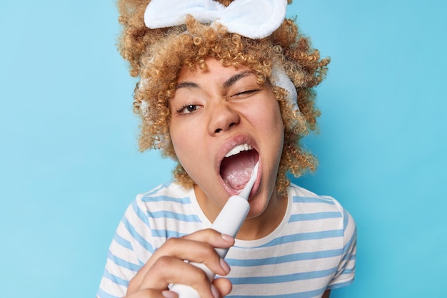 Daily morning routine and brushing habits concept Young female model with curly fair hair brushes teeth with electric toothbrush keeps teeth healthy isolated over blue background Freshness of breath