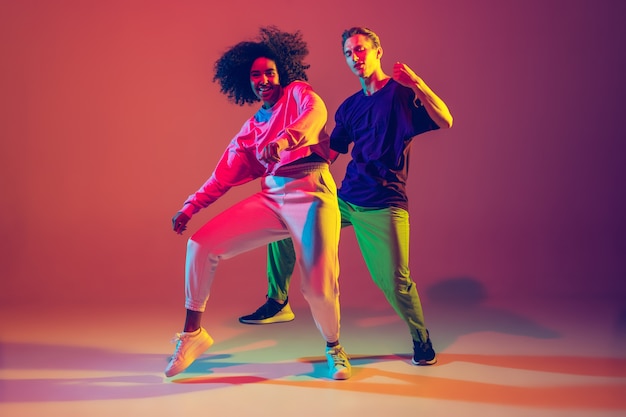 Free Photo dance time. stylish men and woman dancing hip-hop in bright clothes on green background at dance hall in neon light. 