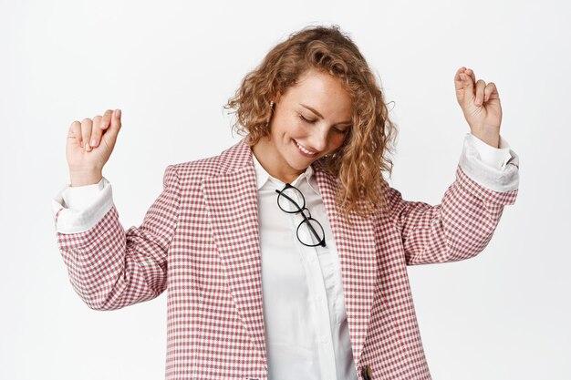 Dancing business woman smiling, raising hands free in air, having fun and relaxing, standing in suit against white background.