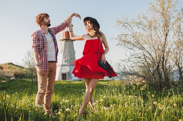 Dancing young stylish couple in love in countryside, indie hipster bohemian style, weekend vacation, summer outfit, red dress, green grass, holding hands