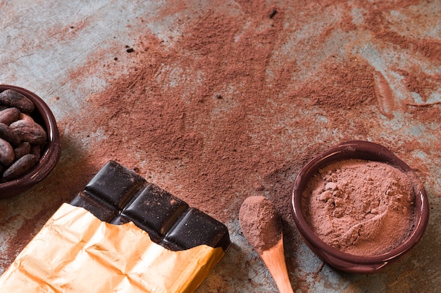 Dark chocolate bar with scattered cocoa powder and beans bowl on rustic backdrop