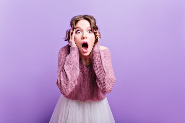 Dark-eyed european woman posing emotionally with mouth open.  surprised brown-haired girl with short haircut touching her head during photoshoot on purple wall.