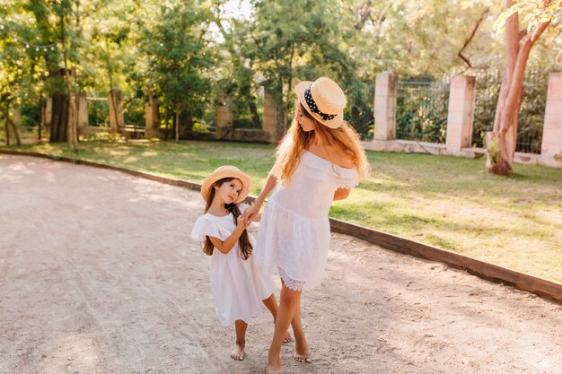 Dark-haired girl begging mother about something looking into her eyes on the street. Outdoor portrait of slim woman in boater holding hands with little daughter.