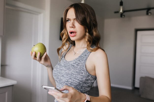 Free photo dark-haired girl with nude makeup posing at home with smartphone. unhappy young woman with apple and phone standing with mouth open.