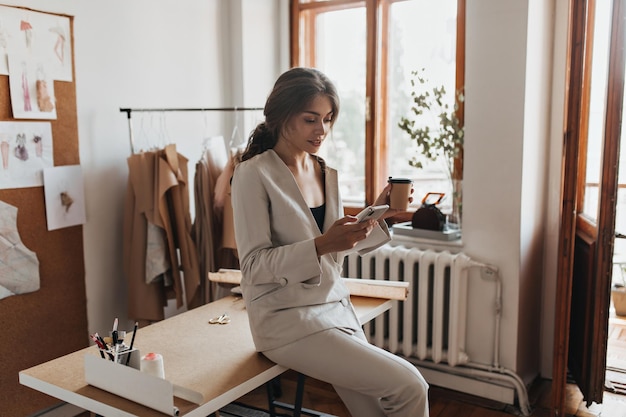 Free photo dark haired lady in white suit holding coffee and phone charming woman in linen pants and beige blouse chatting in cellphone and drinking tea