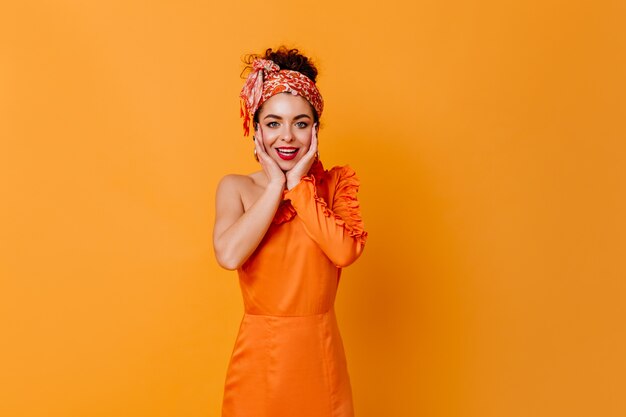 Free photo dark-haired lady with red lipstick dressed in orange dress and aheadband with smile looking at camera on isolated space.