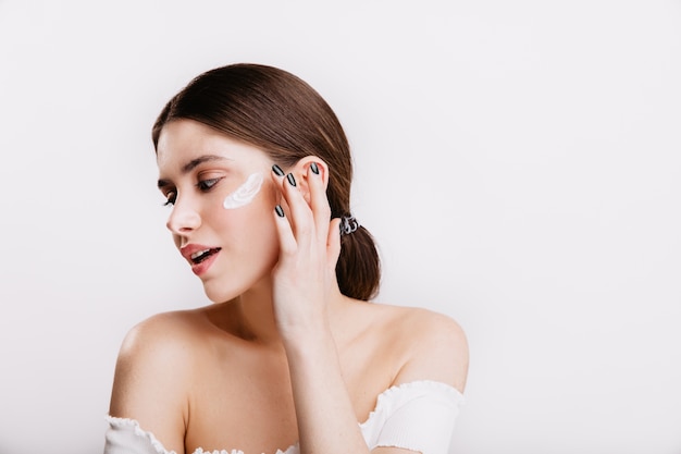 Dark-haired lady without makeup puts moisturizer on her face. Girl with black manicure gently touches skin.