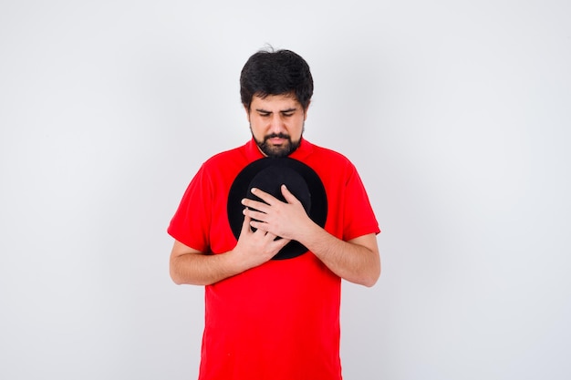 Free photo dark-haired man in red t-shirt hugging his cap and looking hopeful , front view.